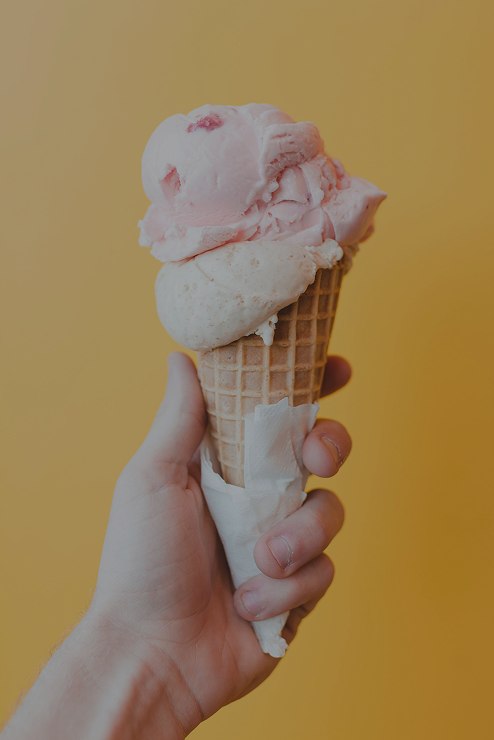 A hand holding a two-scoop ice cream cone against a warm ochre background