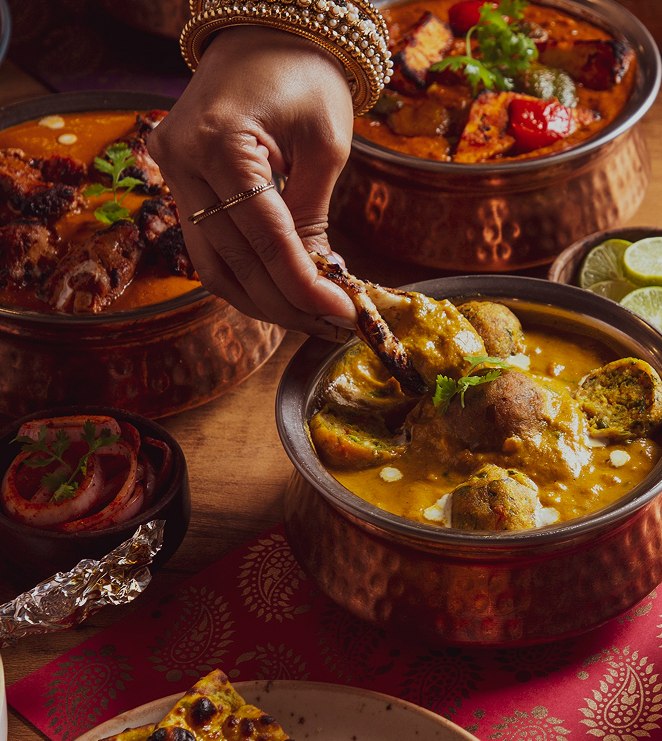 A hand reaching into a spread of curries served in hammered copper bowls on a patterned cloth