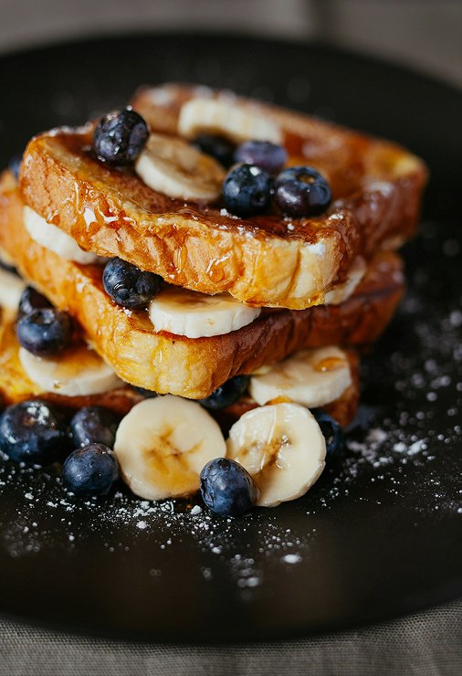 A stack of French toast with banana and blueberries on a dark plate, dusted with sugar